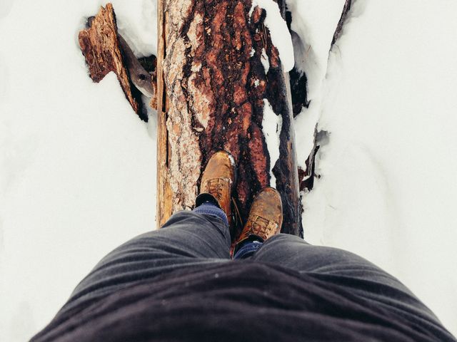 Person balancing on a snow-covered fallen tree trunk during winter. Ideal for promoting outdoor adventure activities, clothing apparel, nature exploration, or travel experiences in cold environments.