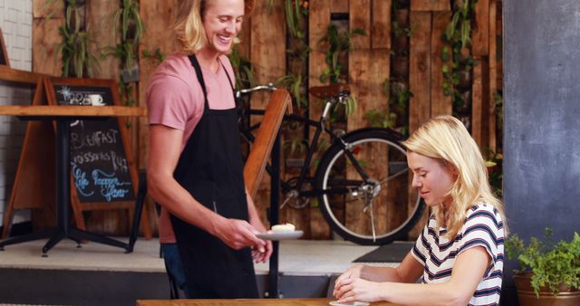 Image shows a smiling blonde waiter serving a dish to a woman with similar hair color in a rustic-themed cafe. Both look happy, showing a friendly and welcoming atmosphere. Useful for depicting customer service, hospitality settings or advertisements for cafes and restaurants.