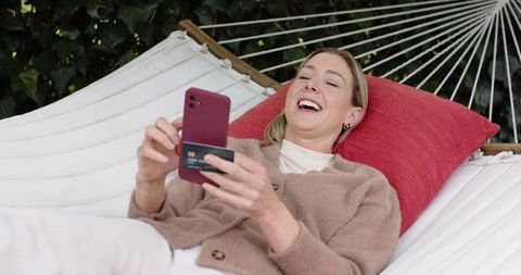 Laughing woman relaxing on hammock using smartphone and credit card in garden