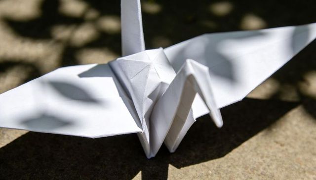 White origami crane resting on sunlit stone patio with leaf shadows, closeup fold crease texture