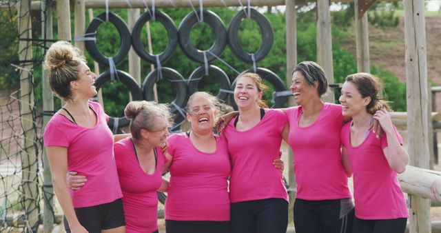 Image showcases a group of women wearing pink shirts, standing together and smiling in an outdoor fitness setting with an obstacle course. This image can be used for promoting team-building activities, outdoor fitness programs, exercise classes, community wellbeing events, or campaigns focused on healthy lifestyles and bonding. Its vibrant and energetic composition can attract audiences interested in group activities and wellness.