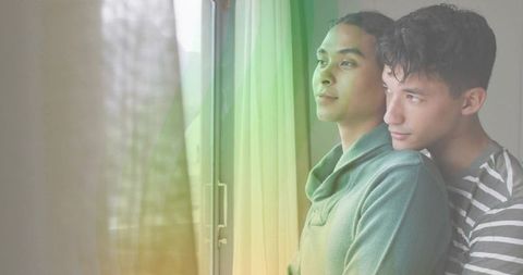 Couple standing by window embracing, gazing outdoors through sheer curtains in daylight