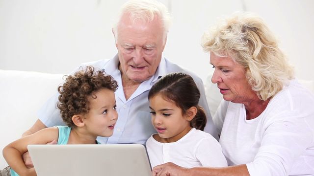 Senior couple and their young grandchildren engaging with a laptop in a bright, cozy living room. This video can be used for articles on family bonding, intergenerational learning, use of technology by seniors, or promotions for tech gadgets geared towards family use.