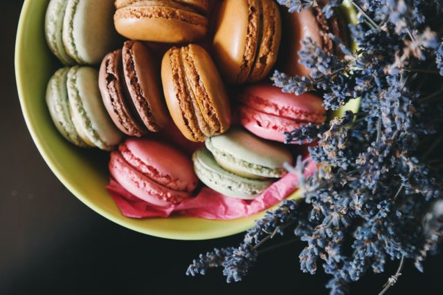 Beautiful assortment of colorful French macarons in green bowl with decorative lavender flowers, exemplifying elegance and taste. Ideal for use in culinary blogs, food magazines, confectionery advertisements, or patisserie menus to showcase gourmet treats.
