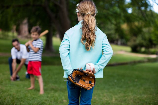 Family enjoying a sunny day playing baseball in a park. Father teaching son to bat while daughter holds glove and ball. Ideal for concepts of family bonding, outdoor activities, summer fun, and childhood memories.