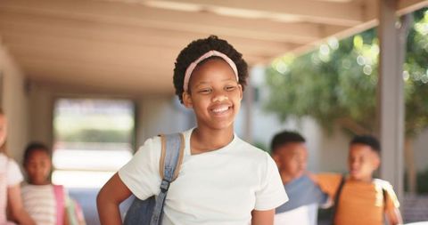 Smiling teenage student walking with denim backpack and classmates on sunny school corridor