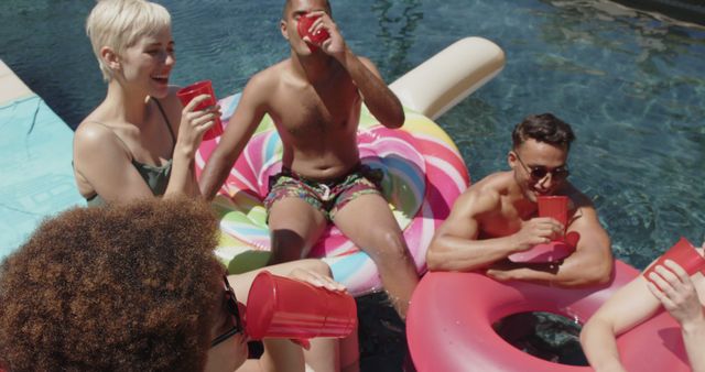Group of young friends enjoying a pool party while lounging on colorful inflatable floats and drinking from red cups on a sunny day. Ideal for use in marketing materials for summer events, promotions for pool-related products, or lifestyle blogs about outdoor fun and social gatherings.