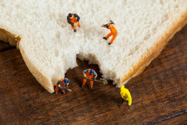 Miniature workers in construction outfits are digging into a slice of bread on a wooden table. This creative and humorous image can be used for advertising, marketing campaigns, or social media posts related to food, creativity, teamwork, or unique concepts. It is ideal for illustrating the idea of hard work and effort in a playful and engaging manner.