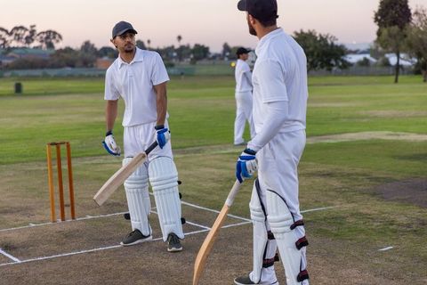 Diverse male cricketers practicing batting at dusk on community pitch for twilight training