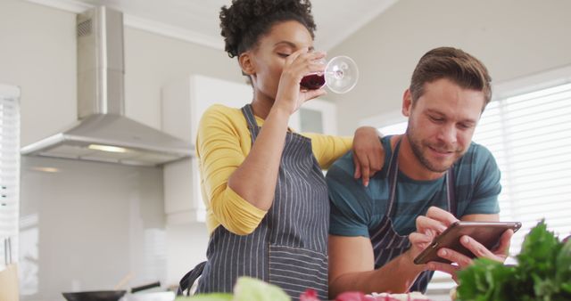 Couple enjoying quality time cooking together in a modern kitchen. Both are wearing aprons, with the woman drinking wine while the man looks at a tablet. Ideal for use in ads related to home appliances, romantic activities, recipe apps, or lifestyle blogs.