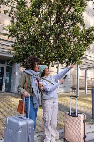 Two women travelers pointing and navigating with smartphone while waiting by luggage in urban plaza