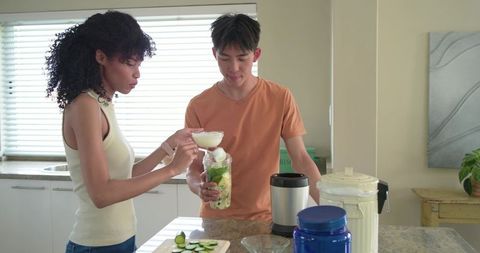 Preparing green cucumber avocado smoothie with protein powder by diverse couple at kitchen island