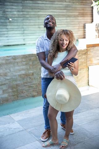 Diverse couple hugging poolside holding straw hat and passport enjoying sunny vacation