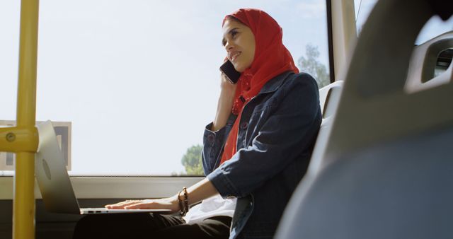 Muslim woman in hijab is commuting on public transport while talking on her mobile phone and using a laptop. This portrays a modern and productive lifestyle. Ideal for use in articles about work-life balance, public transport, or modern women in business.