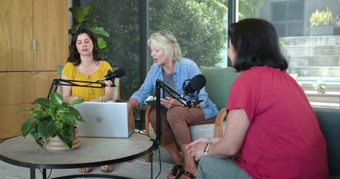 Women hosting podcast roundtable in cozy living room using laptop and microphones