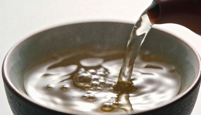 Pouring hot tea into glazed ceramic bowl close-up with steam, ripples and reflections
