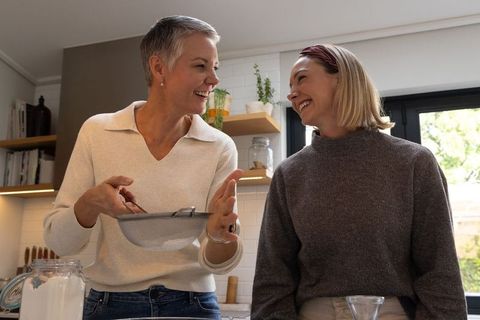 Two women laughing while sifting flour in sunlit home kitchen casual family baking moment