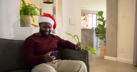 Mid adult African American man relaxing on couch wearing Santa hat and using smartphone