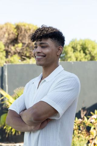 Smiling african american man in white polo standing with arms crossed in sunny backyard