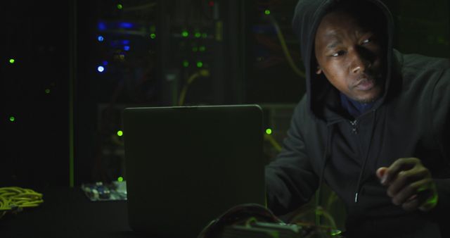 Young man wearing hoodie, seated at table, using laptop in dimly lit data center. Network wires and LED lights visible in the background. Can be used for themes related to cybersecurity, hacking, technology, IT professions, and computer networks.