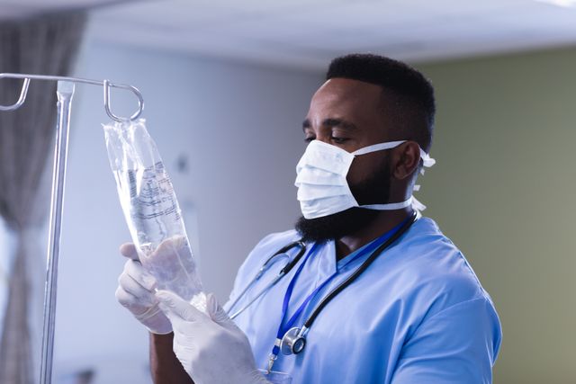 This image depicts an African American male doctor wearing a face mask and gloves, carefully examining an IV drip bag in a hospital setting. The doctor is focused on ensuring the proper administration of fluids or medication to a patient. This image can be used in healthcare-related articles, medical service promotions, hospital websites, and educational materials about patient care and safety protocols.