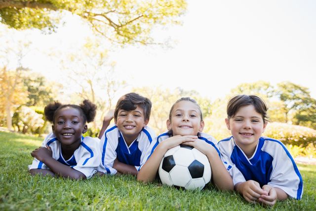 Children soccer team members lying on grass in park, smiling at camera. Ideal for promoting youth sports, teamwork, outdoor activities, and diversity. Suitable for use in advertisements, educational materials, and community event promotions.