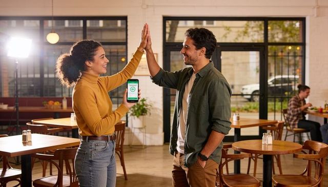High-fiving woman holding smartphone green-check while man celebrates in warm backlit cafe