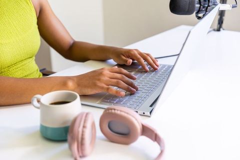 African american woman typing on laptop in home studio with microphone and pink headphones