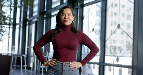 Confident woman standing hands-on-hips in modern glass office, burgundy top and jeans
