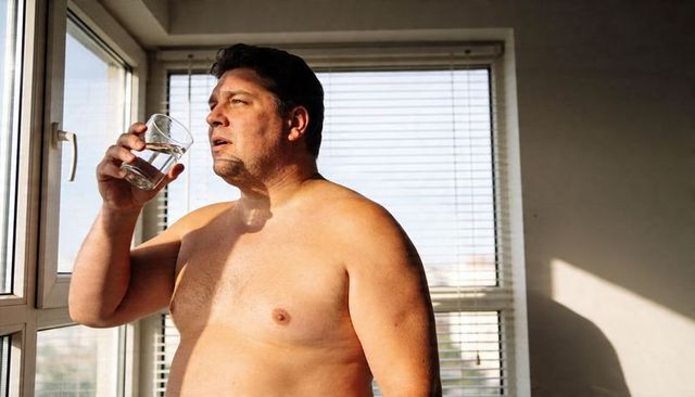 Bare-chested middle-aged man drinking water by sunlit window, contemplative morning mood