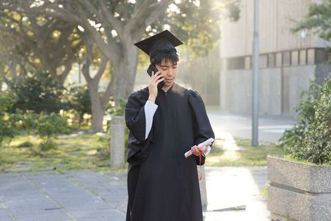 Asian graduate calling on phone while holding diploma in mortarboard and gown sunlit campus