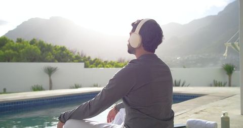 Indian man relaxing poolside with headphones facing sunlit mountains, wellness and leisure