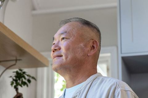 Senior asian man standing and looking upward in bright kitchen with bonsai on shelf
