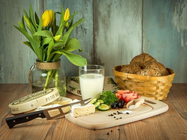 This setting showcases a fresh and nutritious breakfast arrangement on a rustic wooden table. The combination of cheese, bread, and fruits such as strawberries and lime provides a wholesome meal option. The presence of yellow tulips in a glass vase adds a vibrant touch and enhances home decor themes. Perfect for illustrating concepts of healthy eating, nature-inspired meals, cozy kitchen atmospheres, or breakfast recipes in food blogs and magazines.