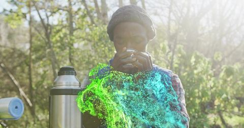 Sipping hot drink in sunlit woods holding cup near thermos with green cyan mist