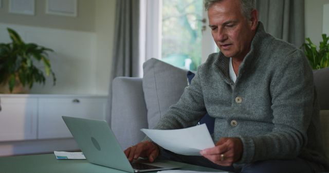 Man in his living room focused on examining documents, suggesting home office or remote work context. Could be used for articles or advertising on remote work benefits, work-life balance, or financial planning. Suitable for illustrating concepts related to telecommuting, office independence, or productivity in a casual home environment.