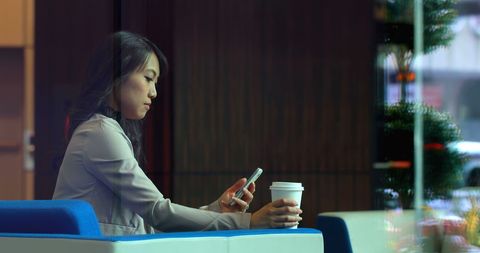 Businesswoman using smartphone while holding coffee cup in modern office