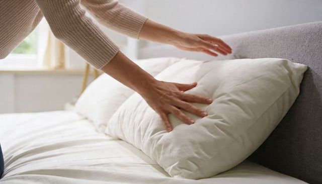Fluffing white pillow on sunlit neutral bed, hands smoothing bedding for cozy bedroom comfort