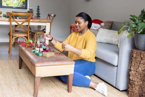 African american woman wrapping holiday gifts in cozy sunlit living room, mustard sweater