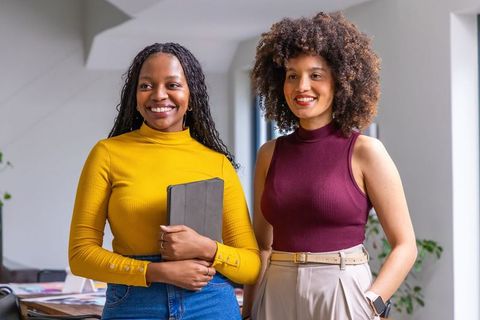 Diverse female coworkers smiling in modern office holding tablet and smartwatch