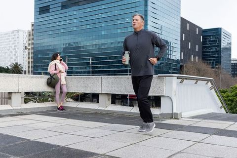 Mature man jogging on urban terrace while woman using smartphone by glass offices