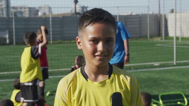 Young boy in a yellow sports jersey standing on a soccer field, holding a microphone. Teammates and coach are seen in the background stretching and talking. Ideal for use in promotions for youth sports programs, team-building activities, sportswear advertisements, or articles on the benefits of youth soccer training.