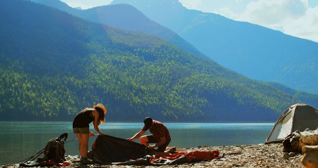 Couple setting up a tent near a picturesque mountain lake on a summer day. Ideal for use in travel blogs, promotional material for outdoor adventure gear, camping trip advertisements, and lifestyle articles focusing on nature activities and relaxation.