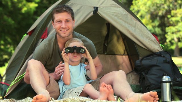 Father and son enjoying camping trip in nature, with child using binoculars under parental guidance. Perfect for family-oriented advertisements, adventure tourism promotions, and outdoor lifestyle content showcasing parent-child bonding.