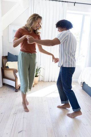 Young couple dancing barefoot on bright wood floor in sunlit bedroom, intimate home moment