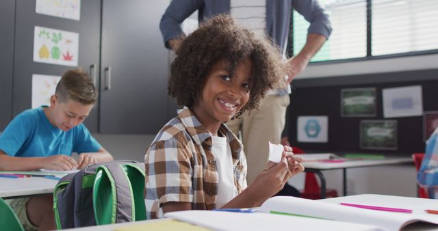 Depicted is a young boy with curly hair smiling while working on a craft project in a classroom. His classmates and a teacher are seen in the background. This would be a useful visual for educational blogs, school brochures, or promotional materials showcasing classroom creativity and student engagement.