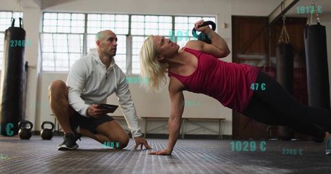 Female athlete balancing kettlebell side-plank while trainer coaching with tablet and ar