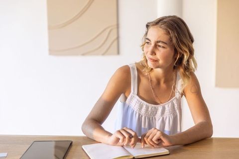 Young woman writing in notebook while gazing at tablet on warm minimalist wooden desk
