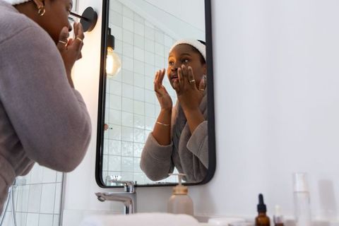 African american woman applying face moisturizer at mirror during skincare routine