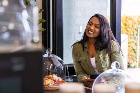 Indian woman smiling at cafe counter holding phone with croissants under cloches